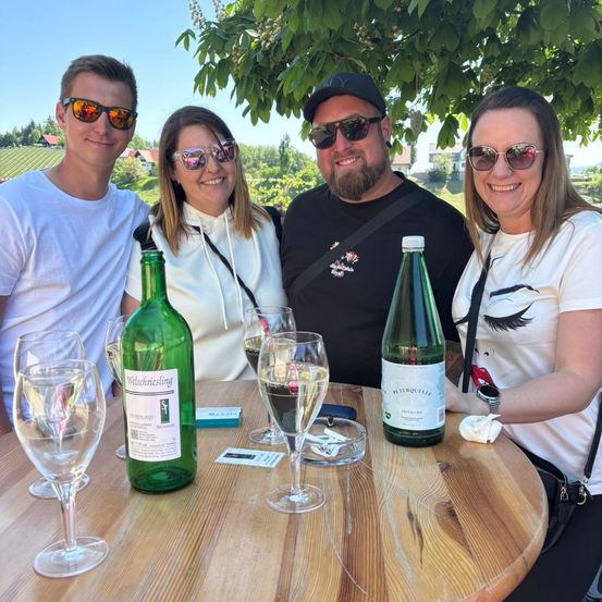 Eine Gruppe von vier Personen sitzt um einen runden Holztisch herum mit Wein-gläsern, Weinflaschen und einem Baum im Hintergrund.