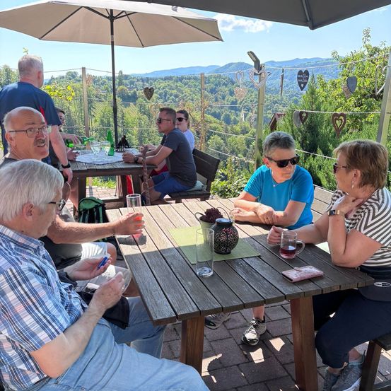 Gruppe von Menschen sitzt um einen Tisch auf einer Terrasse und genießt Getränke mit einem malerischen Blick auf die Berge im Hintergrund.