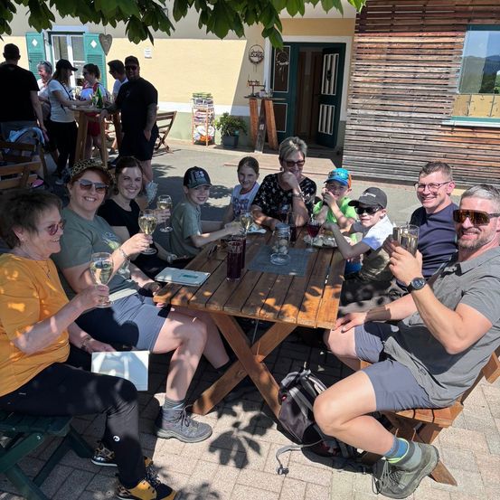 Eine Gruppe von Erwachsenen und Kindern sitzt draußen um einen Holztisch und hält Weinglas in der Hand und lächelt, mit einem Gebäude und einem Baum im Hintergrund.