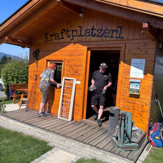 Zwei Personen stehen vor einer Kraftplatzerl-Hütte mit einer Speisekarte und einem Müllcontainer. Eine Frau hält eine Speisekarte und ein Mann betritt die Hütte.