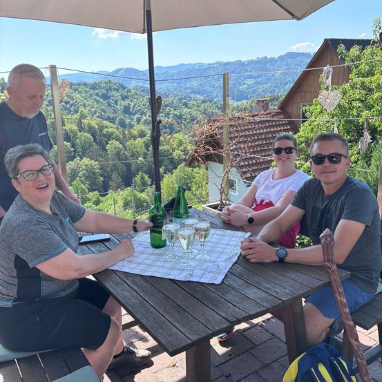 Drei Personen sitzen um einen Tisch auf einem Balkon. Sie lächeln und scheinen eine Mahlzeit zu genießen. Eine Flasche Wein und Weingläser stehen auf dem Tisch. Die Frau trägt eine Brille.