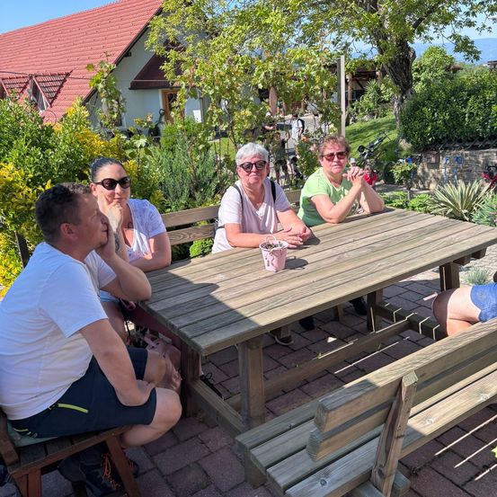 Eine Gruppe von Menschen sitzt um einen Holztisch in einem Garten, wobei zwei Frauen eine Sonnenbrille tragen und ein Mann seinen Mund mit der Hand bedeckt.