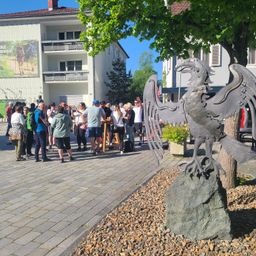 Eine Gruppe von Menschen versammelt sich um eine große Metall-Adlerstatue auf einem Felsen in einer Stadtmitte. Ein Gebäude mit einem Schild und einem Baum steht hinter ihnen.