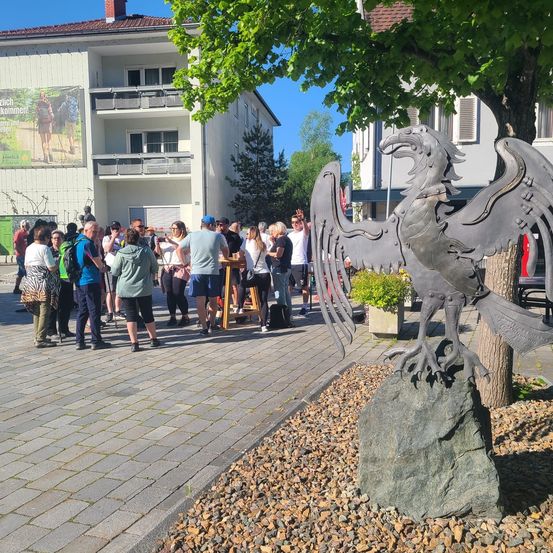Eine Gruppe von Menschen versammelt sich um eine große Metall-Adlerstatue auf einem Felsen in einer Stadtmitte. Ein Gebäude mit einem Schild und einem Baum steht hinter ihnen.