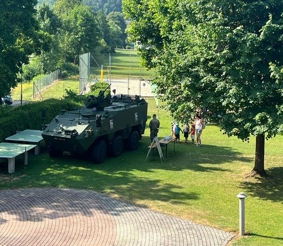 Ein gepanzertes Militärfahrzeug steht auf einem Rasen mit einem Tisch und Stühlen. Menschen gehen in der Nähe. Es gibt auch geparkte Autos und einen Zaun auf der Seite.