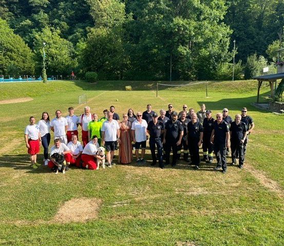 Eine Gruppe von Menschen, darunter einige in Uniform, posiert für ein Foto auf einer Rasenfläche mit Bäumen und einem Gebäude im Hintergrund. Einige Personen halten Hunde.