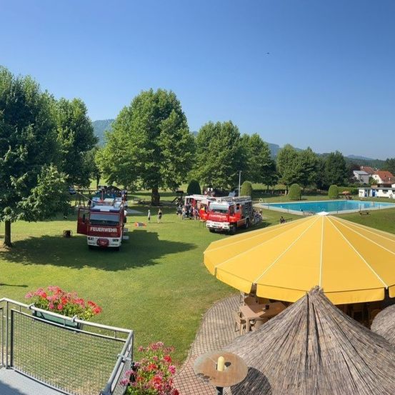 Ein Blick von oben auf einen Park mit Pool, Bäumen und Menschen. Zwei Feuerwehrwagen sind in der Nähe des Pools geparkt, mit Leuten, die herumstehen. Ein gelber Regenschirm befindet sich in der Nähe, und ein Zaun umgibt das Gebiet.