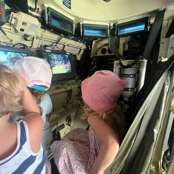 Drei Kinder sitzen in einem Militärfahrzeug. Ein Kind trägt eine rosa Mütze und schaut auf einen Monitor. Ein weiteres Kind mit einer weißen Mütze schaut nach oben. Das Fahrzeug hat mehrere Monitore und Kabel.