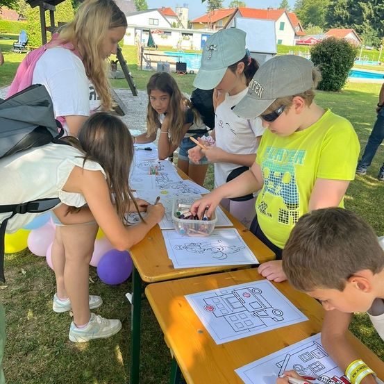 Kinder zeichnen ein Bild auf einem Tisch. Zwei Mädchen stehen. Eines hält einen Marker. Im Hintergrund ist ein Schwimmbad.