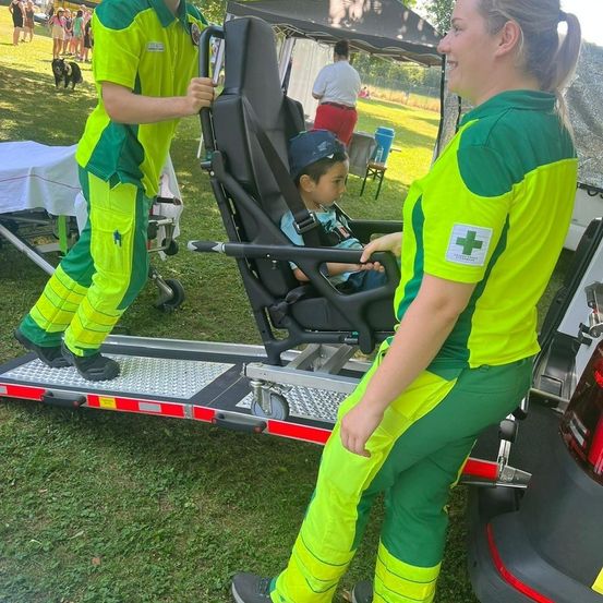 Zwei Personen in grünen Uniformen helfen einem kleinen Kind in einem Stuhl aus einem Krankenwagen. Das Kind trägt eine blaue Mütze. Im Hintergrund befindet sich ein Zelt und ein Tisch mit einem blauen Fass.