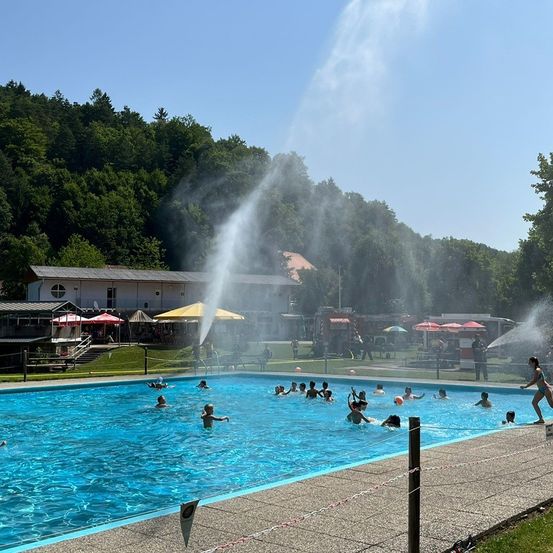 Ein großer Außenpool mit mehreren schwimmenden Personen. Ein Wasserbrunnen sprüht Wasser in die Luft. Im Hintergrund befinden sich Schirme und ein Gebäude.