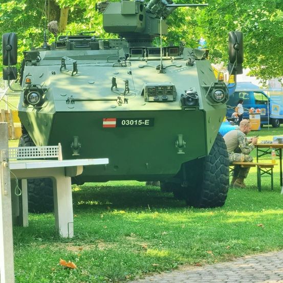 Ein Militärfahrzeug ist auf einem Rasen geparkt. Ein Mann in Tarnkleidung sitzt in der Nähe und isst. Bäume und ein Lieferwagen sind im Hintergrund.