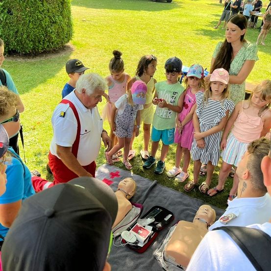 Ein Mann mit roten Hosenträger zeigt einer Gruppe von Kindern wie man einen CPR-Kurs mit einer Puppe auf einer Decke durchführt. Andere Erwachsene und Kinder beobachten im Park.
