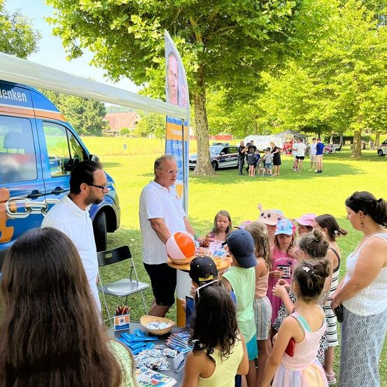 Eine Gruppe von Kindern und Erwachsenen versammelt sich um einen Tisch in einem Park, mit einem Polizeiwagen und einem Auto im Hintergrund. Ein Mann spricht zu den Kindern.