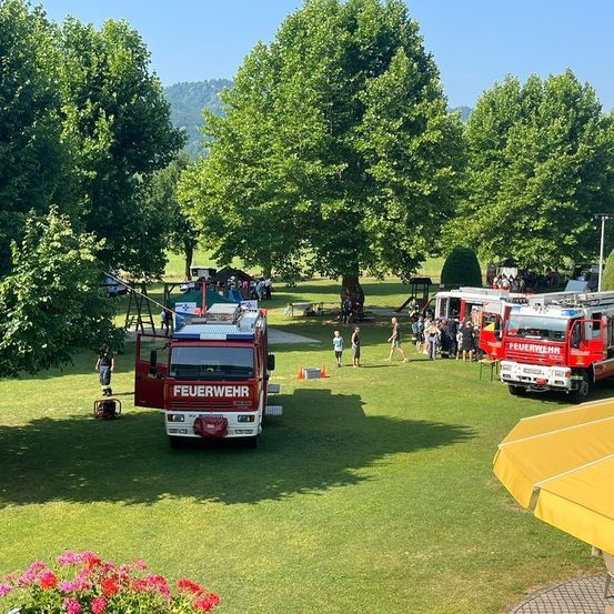 Ein Feuerwehrauto ist auf einem Rasen geparkt, mit mehreren Menschen und anderen Fahrzeugen in der Nähe. Es gibt Bäume und ein Zelt im Hintergrund.
