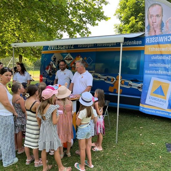 Eine Gruppe von Kindern und Erwachsenen versammelt sich um ein Zivilschutzfahrzeug und hört einem Mann in einem weißen Hemd zu. Hinter dem Fahrzeug ist ein Banner mit dem Gesicht eines Mannes angebracht.