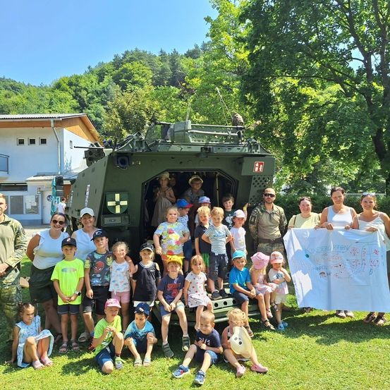 Eine Gruppe von Kindern und Erwachsenen posiert für ein Foto vor einem Militärfahrzeug in einem Park mit Bäumen und einem Gebäude im Hintergrund.