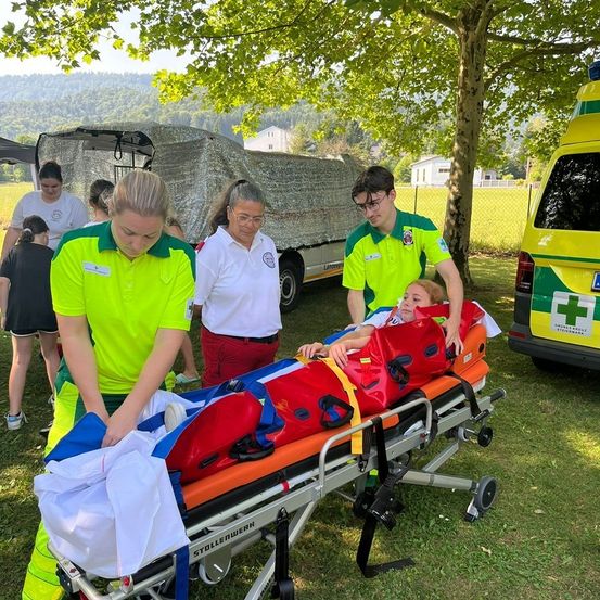Freiwillige in grünen und gelben Uniformen helfen einer Person auf einer Trage unter einem Baum. Dahinter ist ein Krankenwagen geparkt. Der Ort ist im Freien mit einem Zaun und Bäumen.