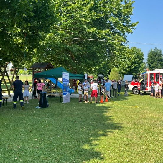 Eine sonnige Parkszene mit einem grünen Zelt, einem Feuerwehrauto und verschiedenen Personen, die sich an Aktivitäten beteiligen, einschließlich einer Reihe von Kindern.