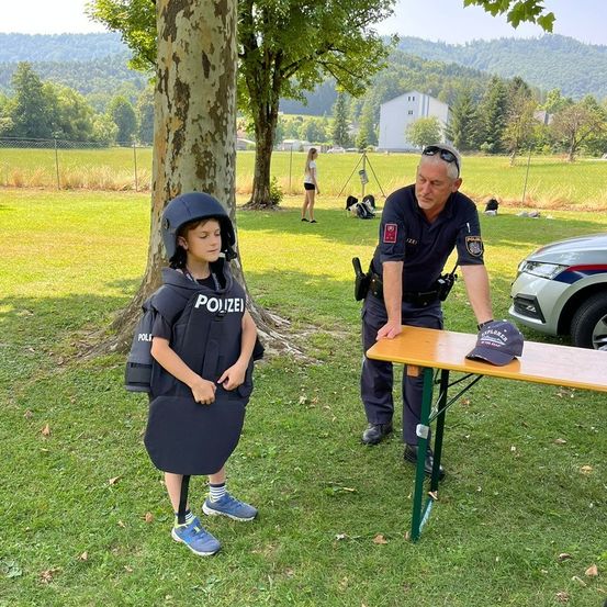 Ein Polizeibeamter steht neben einem Kind in einer Polizeiweste auf einem Rasenfeld, in der Nähe eines Tisches mit einer Mütze. Im Hintergrund befinden sich ein Zaun, eine Frau und ein weißes Gebäude.