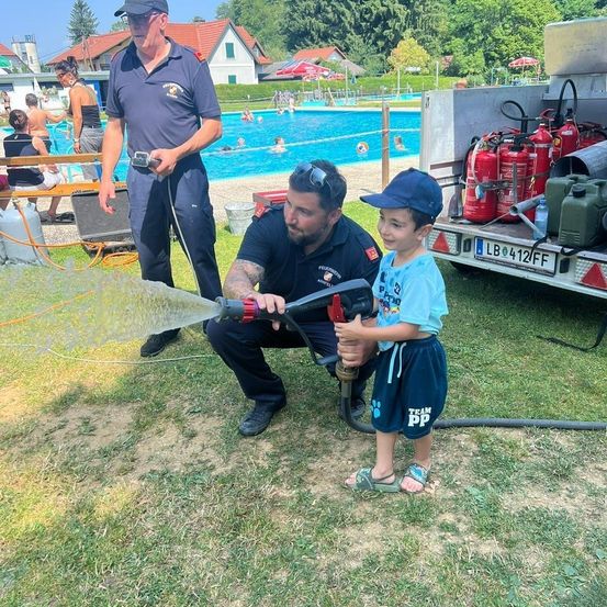 Zwei Feuerwehrleute zeigen einem kleinen Jungen, wie man einen Wasserschlauch benutzt, in der Nähe eines Schwimmbeckens, mit einem Feuerwehrwagen im Hintergrund.