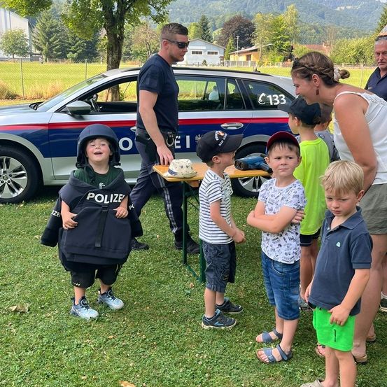 Eine Gruppe von Kindern mit einer Frau und einem Polizeibeamten auf einem Feld. Ein Kind trägt eine Polizeiweste und einen Helm. Ein Polizeiauto ist hinter ihnen geparkt.