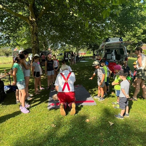Eine Gruppe von Kindern und Erwachsenen versammelt sich in einem Park unter einem Baum. Eine Person in suspenders kniet auf einer Matte mit einer Puppe. Hinter ihnen sind zwei Fahrzeuge geparkt. Einige Kinder tragen Hüte und Schuhe. Ein Tisch mit einer Flasche befindet sich auf der linken Seite.