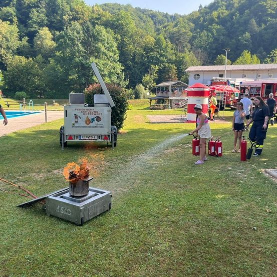 Eine Frau löscht ein Feuer mit einem Feuerlöscher, während Menschen zusehen. Es gibt rote Feuerlöscher und ein Löschtrainingsfahrzeug auf dem Rasen.