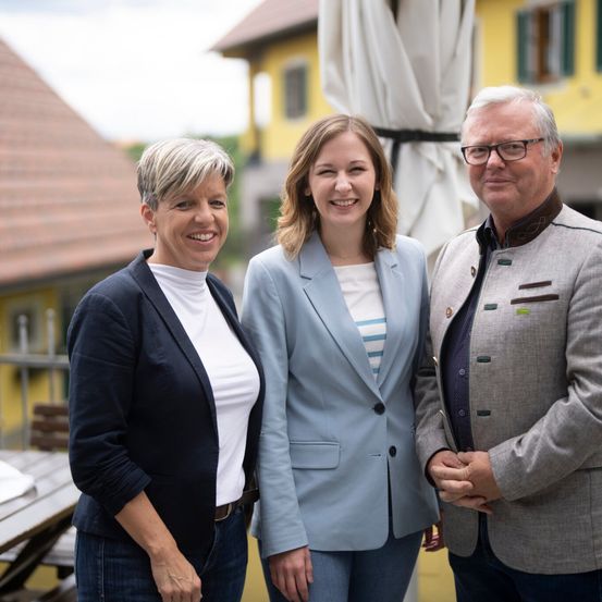 Drei Personen stehen vor einem gelben Gebäude und lächeln. Eine Frau in einer blauen Blazerjacke, eine Frau in einer marineblauen Blazerjacke und ein Mann in einem grauen Jackett.