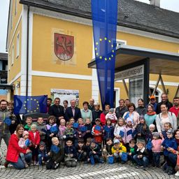 Eine Gruppe von Menschen steht vor einem Gebäude mit einer europäischen Flagge und hält EU-Fahnen. Einige tragen Mützen und Jacken, ein Schild mit der Aufschrift 'MARKTGEMEINDEATM'. Kinder in der Mitte halten EU-Fahnen.