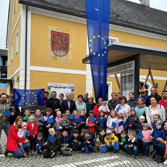 Eine Gruppe von Menschen steht vor einem Gebäude mit einer europäischen Flagge und hält EU-Fahnen. Einige tragen Mützen und Jacken, ein Schild mit der Aufschrift 'MARKTGEMEINDEATM'. Kinder in der Mitte halten EU-Fahnen.