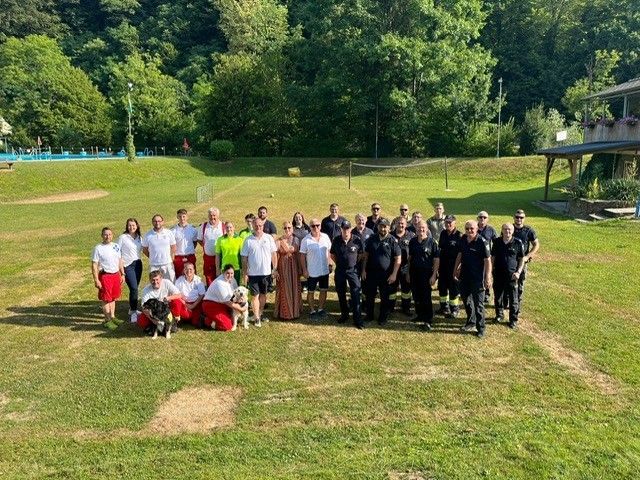 Eine Gruppe von Menschen, darunter Feuerwehrleute und Freiwillige, posiert sonnig für ein Foto mit Hunden und einem Pool im Hintergrund.