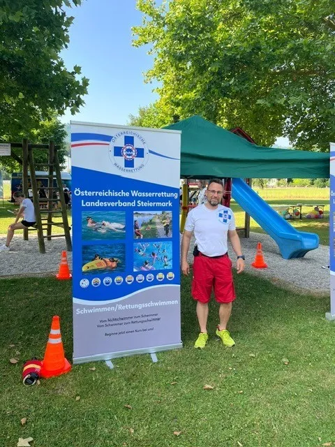 Ein Mann steht in einem Park mit einem Banner des Osterreichischen Wasserrettungsverbandes. Hinter ihm sind eine Rutsche und ein Zelt zu sehen.