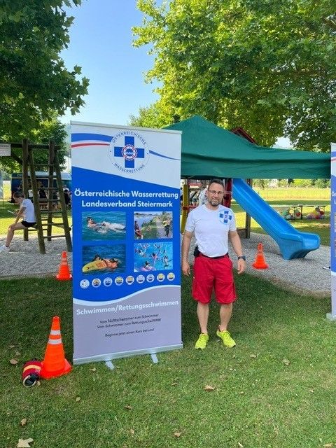 Ein Mann steht in einem Park mit einem Banner des Osterreichischen Wasserrettungsverbandes. Hinter ihm sind eine Rutsche und ein Zelt zu sehen.