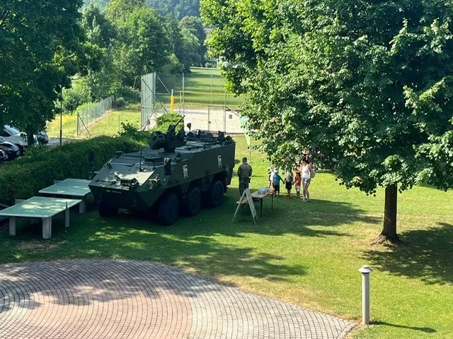 Ein gepanzertes Militärfahrzeug steht in einem Park mit einem Tisch und Bänken, umgeben von Menschen und Bäumen.