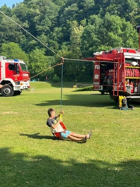 Ein Junge liegt auf einer Schaukel, die an einem Pfahl befestigt ist, und hält sich an der Leine fest. Dahinter steht ein Feuerwehrauto.