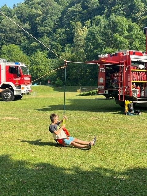 Ein Junge liegt auf einer Schaukel, die an einem Pfahl befestigt ist, und hält sich an der Leine fest. Dahinter steht ein Feuerwehrauto.