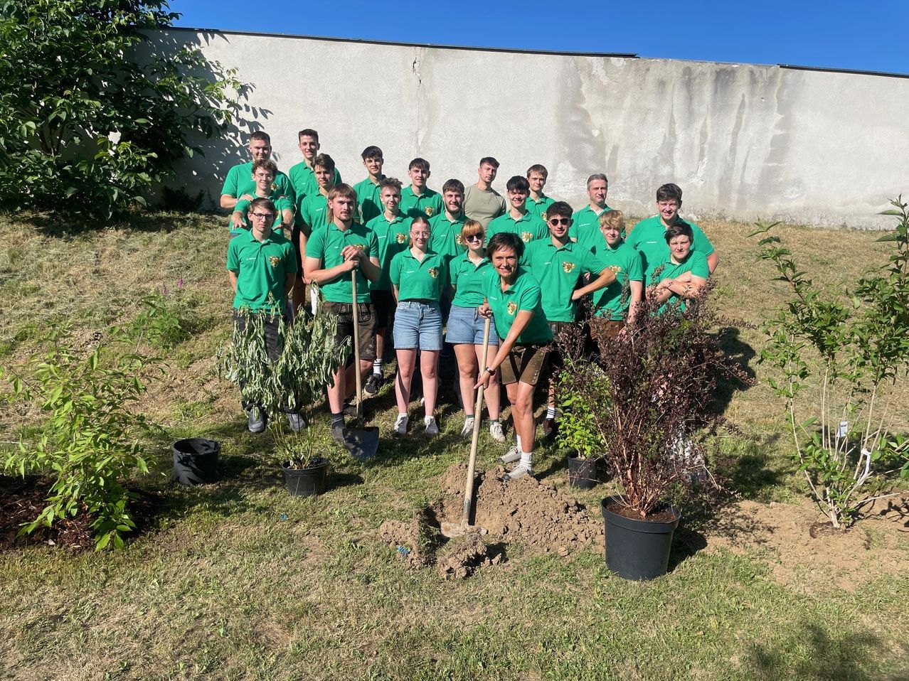 Eine Gruppe von Menschen in grünen Uniformen pflanzt Sträucher in einem Garten. Sie stehen hinter einer weißen Mauer mit einigen Topfpflanzen.
