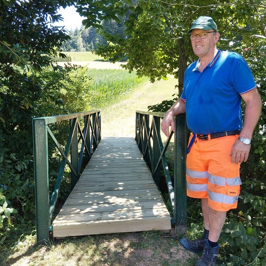 Ein Mann in einem blauen Shirt und orange Shorts steht auf einer hölzernen Brücke mit grünen Geländern, umgeben von Grün.