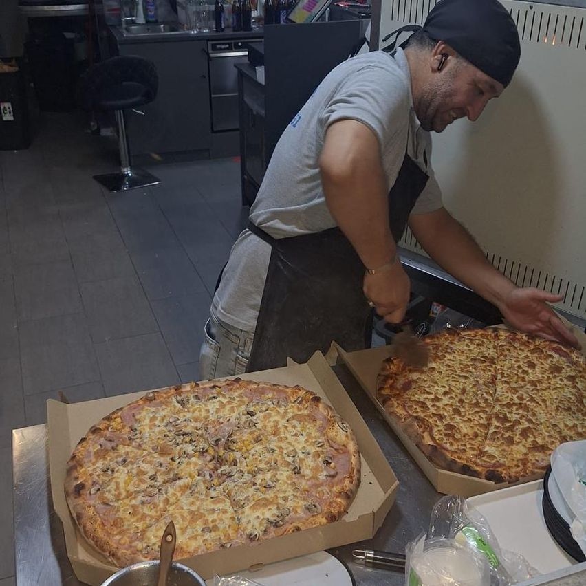 Ein Mann in einem grauen Shirt und einer schwarzen Schürze bereitet große Pizzen mit verschiedenen Belägen in einer Küche zu. Die Pizzen stehen auf einem Tresen mit einer Schüssel, einem Löffel und Tellern.