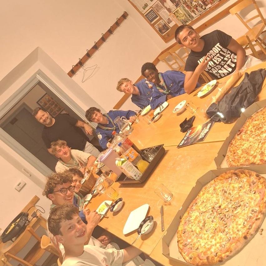 Eine Gruppe von Jungen und Männern sitzt um einen Esstisch und genießt Pizza. Zwei große Pizzen sind auf dem Tisch, zusammen mit verschiedenen Getränken und Besteck. Ein gerahmtes Bild hängt an der Wand.