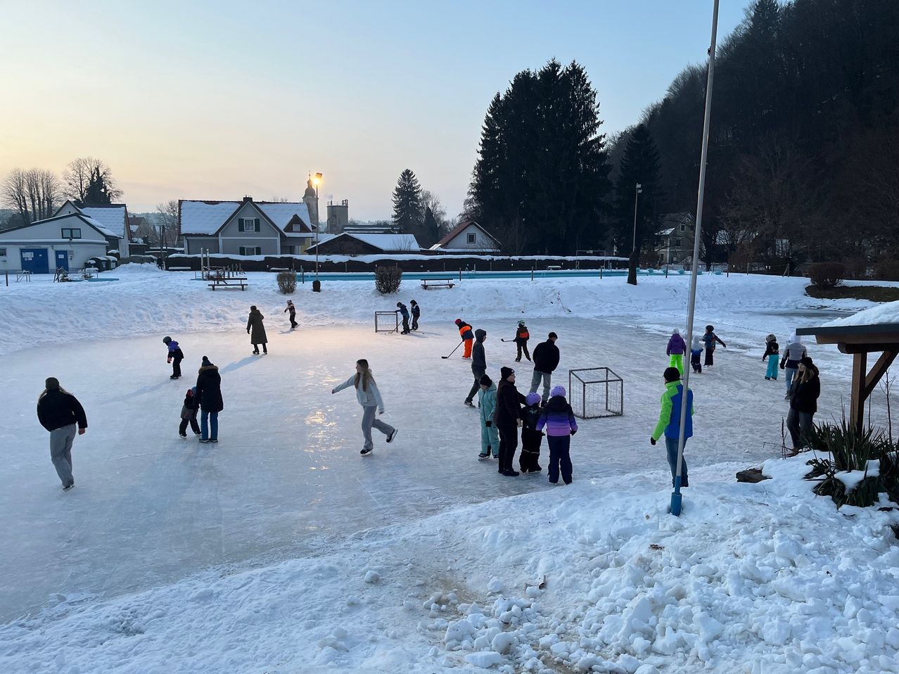 Eine Gruppe von Menschen, die auf einer gefrorenen Eisbahn Schlittschuh laufen, umgeben von Schnee und Häusern im Hintergrund.