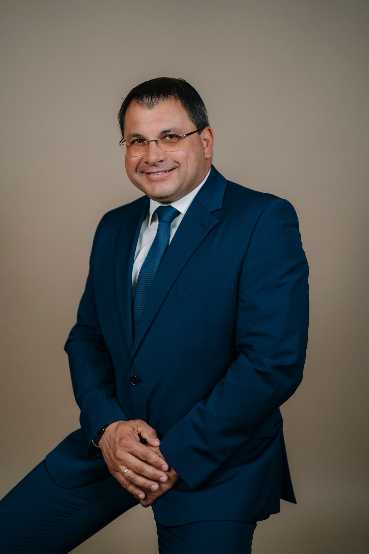 Ein Mann in einem blauen Anzug und Krawatte, mit einer Brille, lächelt, während er gegen einen beigen Hintergrund sitzt.