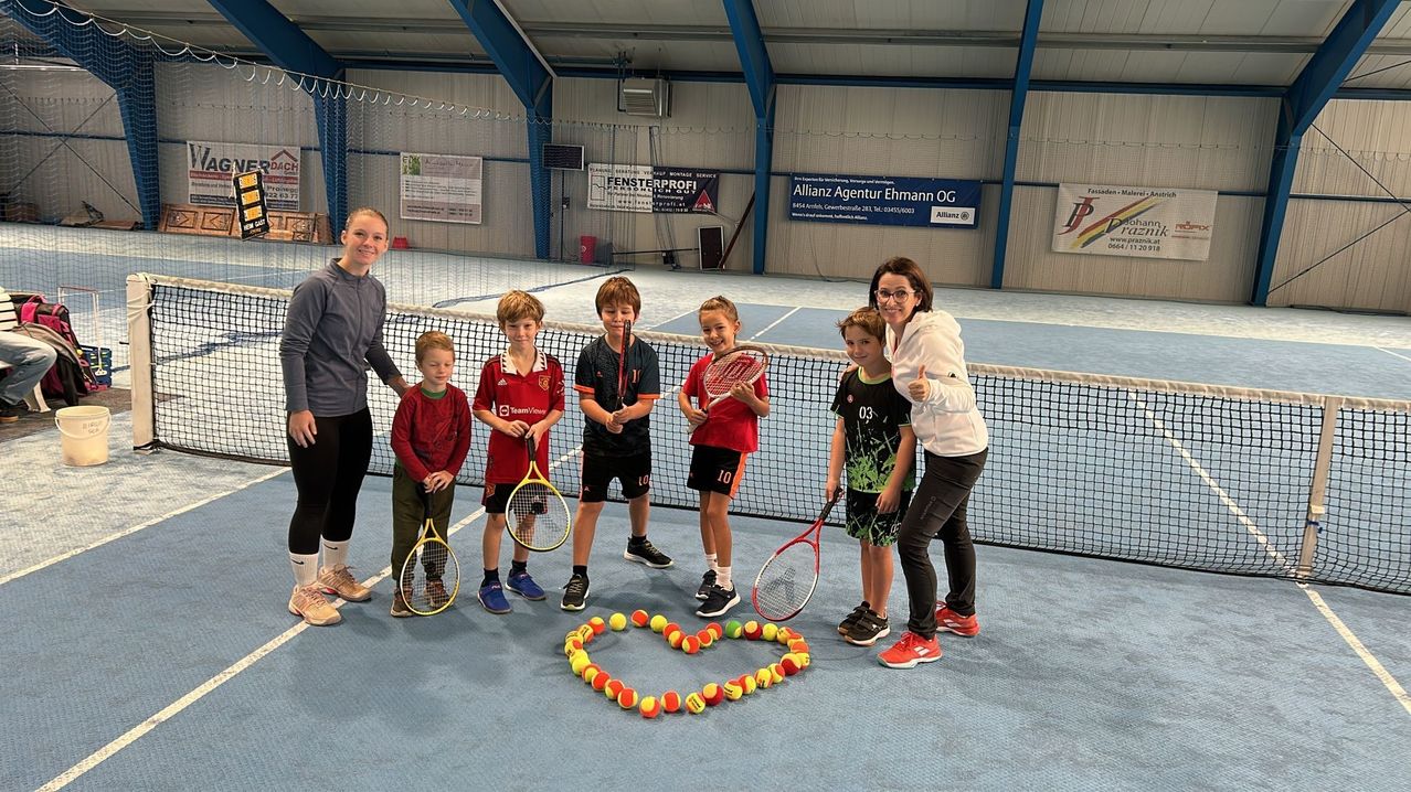 Eine Gruppe von Kindern und zwei Erwachsenen steht auf einem Tennisplatz und posiert für ein Foto mit Tennisschlägern. Tennisbälle sind in Herzform auf dem Boden angeordnet.