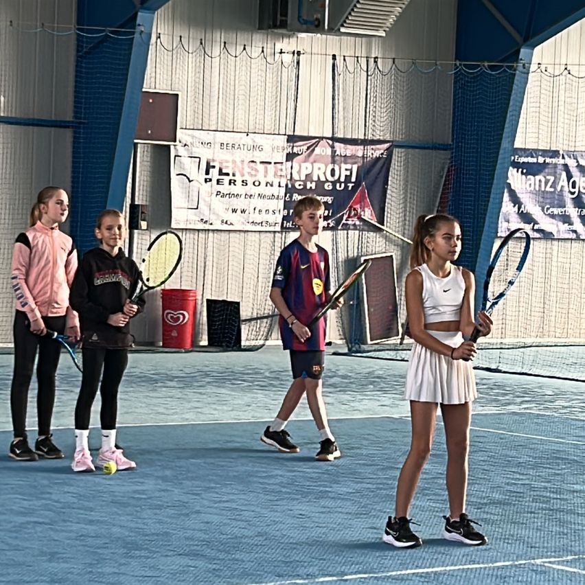 Vier junge Tennisspieler stehen auf einem blauen Tennisplatz. Sie halten Tennisschläger, und ein Ball liegt auf dem Boden. Plakate befinden sich an den Wänden hinter ihnen.