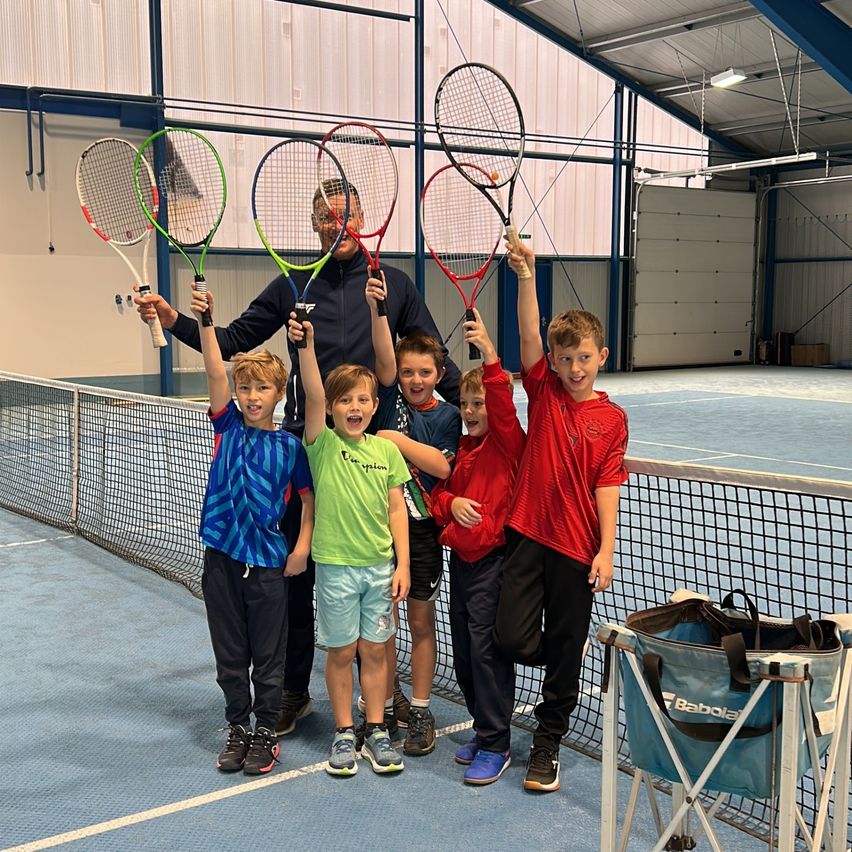 Eine Gruppe von Kindern und einem erwachsenen Mann, die Tennisschläger halten, stehen auf einem Tennisplatz mit einem Netz und einer blauen Tasche mit dem Wort 'BABOLA'.