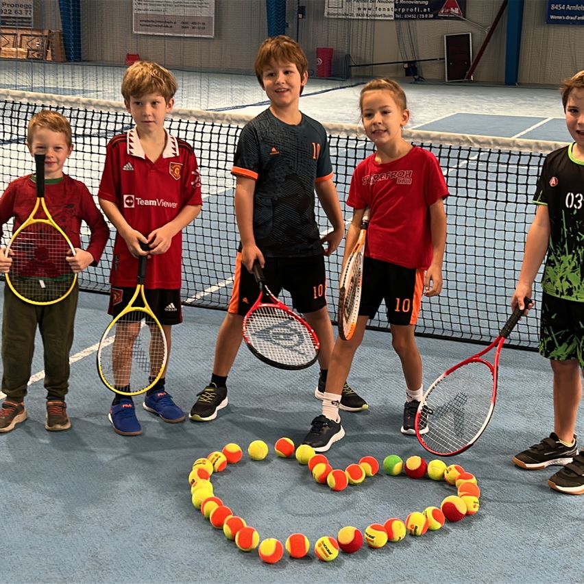 Fünf Jungen in roten und schwarzen Trikots, die Tennisschläger halten, stehen auf einem Tennisplatz. Sie sind von einer herzförmigen Anordnung von Tennisbällen umgeben. Dahinter befindet sich ein Netz und ein Tennisplatz.