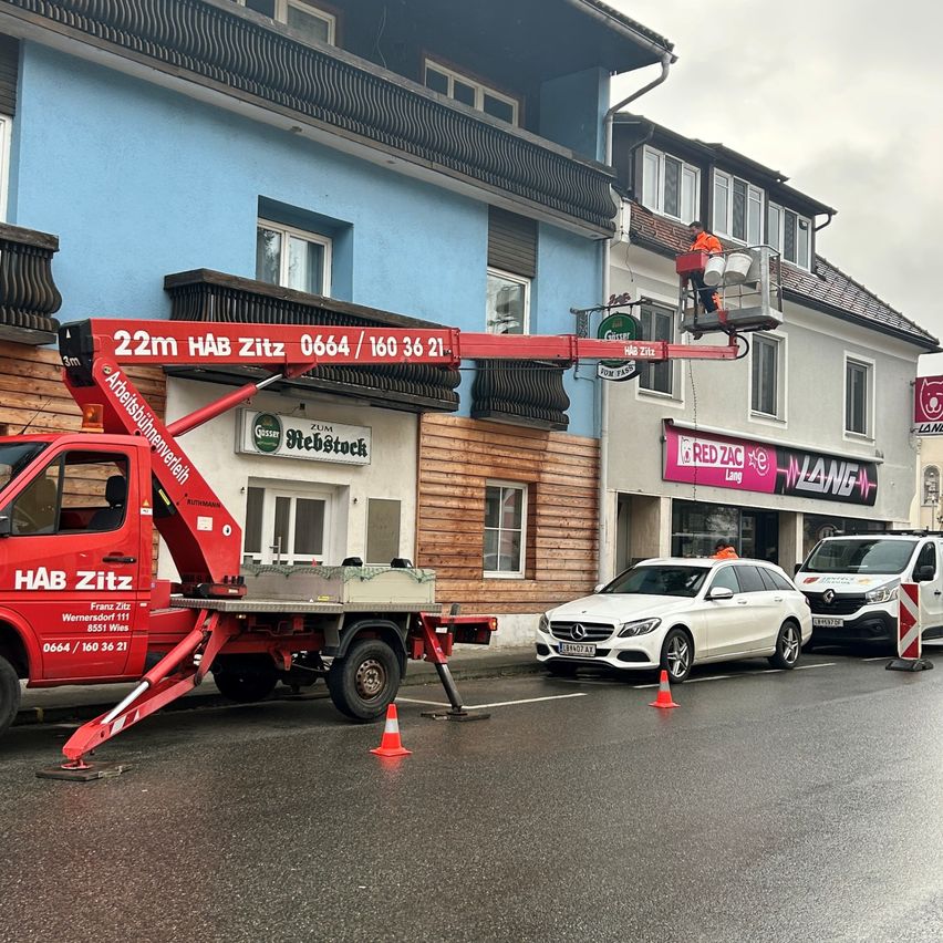 Ein roter Kranwagen steht am Straßenrand, mit einem Arbeiter auf dem Kran. Das Gebäude dahinter hat einen Balkon und Fenster. Ein weißes Auto ist in der Nähe geparkt.