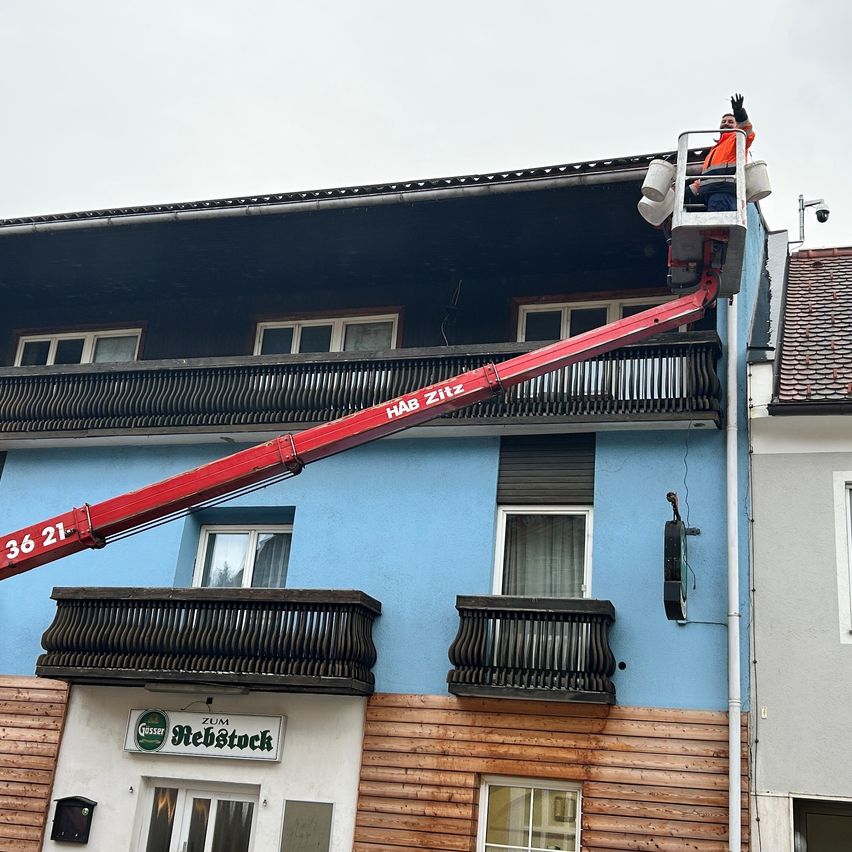 Eine Person benutzt einen roten Kran, um die blaue Wand eines Gebäudes zu streichen. Der Kran ist mit 'HAB Ziltz' beschriftet. Das Gebäude hat Holzverkleidungen und ein Schild mit der Aufschrift 'Zum Rebstock'. Der Himmel ist bedeckt.
