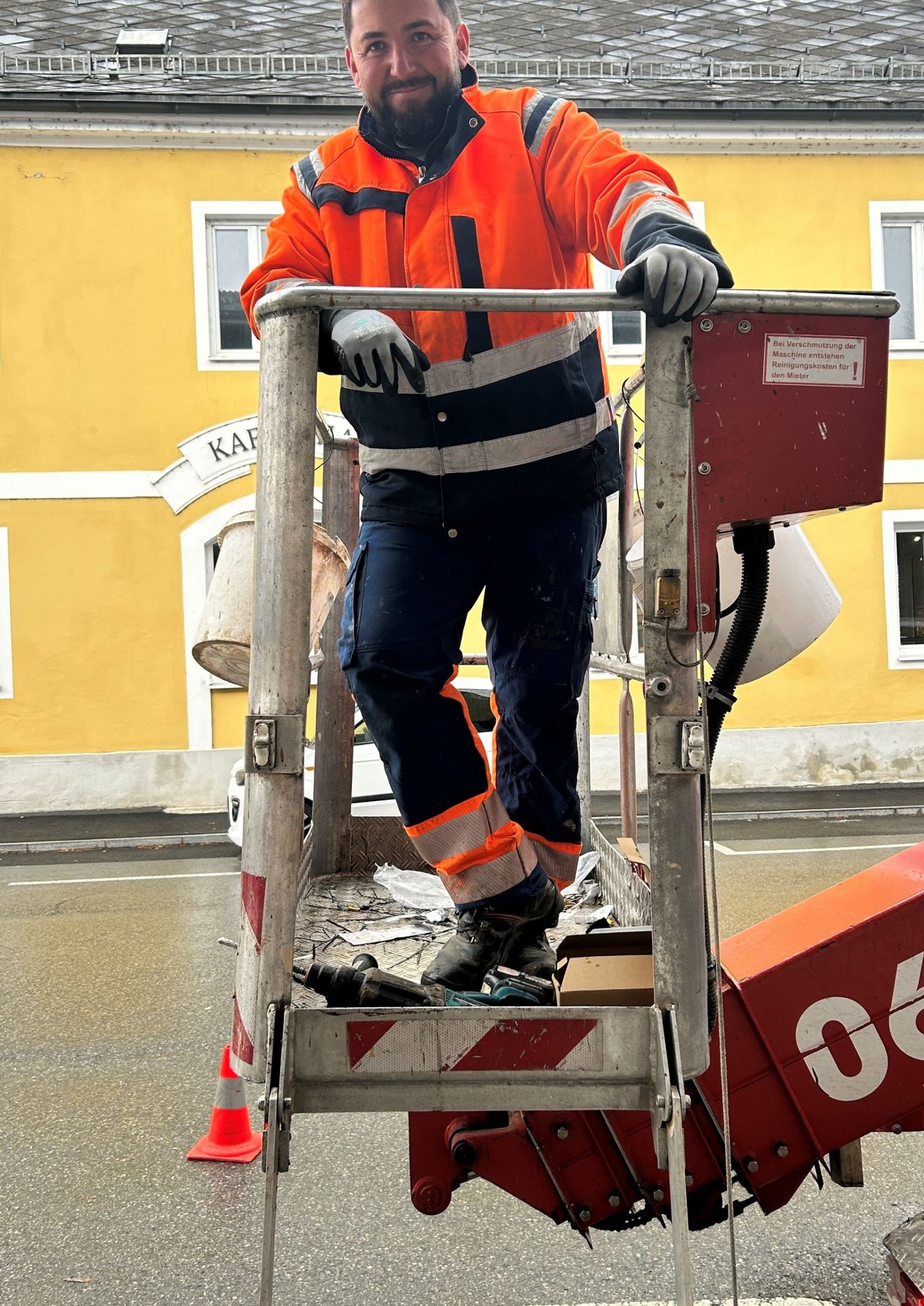 Eine Person in einem orangefarbenen und schwarzen Arbeitsanzug steht auf einer Leiter vor einem Gebäude. Die Leiter ist rot und weiß.